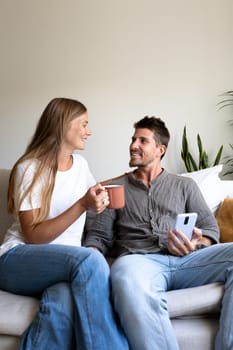 Vertical portrait of happy couple enjoying morning time together drinking coffee and relaxing sitting on the couch. Lifestyle concept.