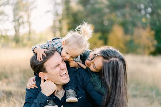 Laughing dad is sitting on the lawn with a little girl on his shoulders and mom hugging them from behind. High quality photo