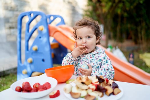 Little girl eats while standing in front of a set table with cut fruits on plates in the garden. High quality photo