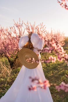 Woman blooming peach orchard. Against the backdrop of a picturesque peach orchard, a woman in a long white dress and hat enjoys a peaceful walk in the park, surrounded by the beauty of nature