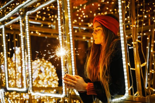 Woman holding sparkler night while celebrating Christmas outside. Dressed in a fur coat and a red headband. Blurred christmas decorations in the background. Selective focus.