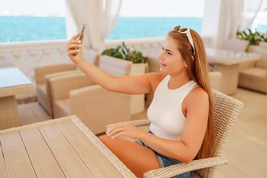 A woman is sitting at a table with a cell phone in her hand. She is taking a picture of the ocean