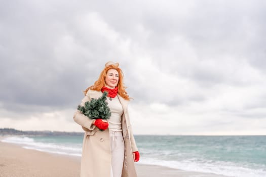 Redhead woman Christmas tree sea. Christmas portrait of a happy redhead woman walking along the beach and holding a Christmas tree in her hands. Dressed in a light coat, white suit and red mittens