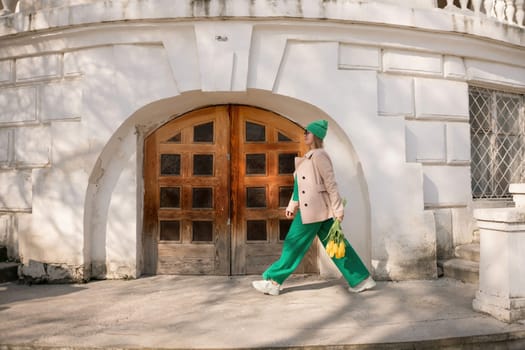 A woman in a green suit and green pants is walking down a street with a bouquet of yellow flowers in her hand