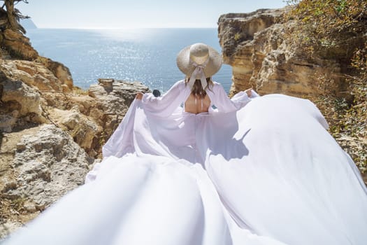 A woman in a white dress is standing on a rocky beach with her hat on. The scene is serene and peaceful, with the woman enjoying the view of the water.