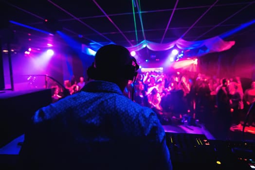 silhouette of a DJ from back in the headphones playing in a nightclub with a crowd of people on the dance floor with multicolored light