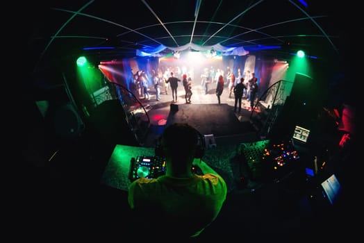 DJ in the night club mixing music on a mixer with headphones and people dancing on the dance floor with bright spotlights