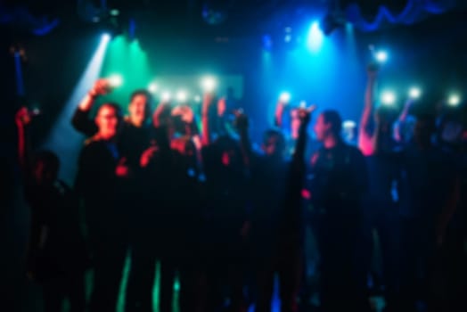 blurred silhouettes of people with flashlights in hand at the concert on the dance floor under the colorful background spotlight