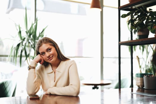 A woman is sitting at a table with her hand resting on her head. She appears contemplative or stressed, deep in thought or experiencing a headache. The setting is indoors, with typical furniture and lighting.