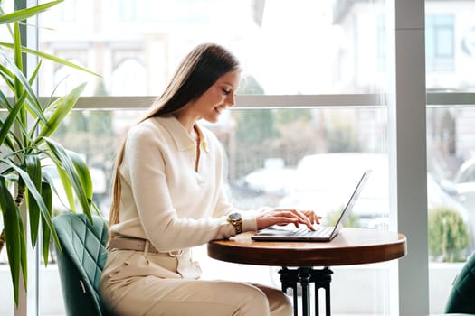 A cheerful woman stands at a high table in a modern cafe, intently focused on her laptop. Natural light floods the space, highlighting both the indoor greenery and the bustling street visible through the large window beside her. The cozy ambiance suggests a productive yet relaxed work environment.