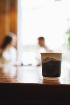 A close-up view of a coffee cup takes the center stage on a wooden table with a blurred background where two people appear to be engaging in a casual conversation in a cozy, indoor setting.