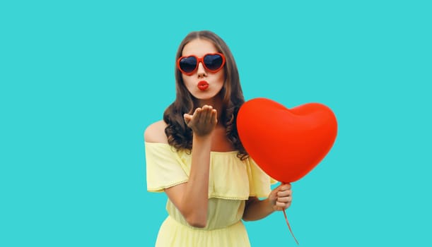 Portrait of happy young woman with red heart shaped balloon blowing kiss in sunglasses on blue studio background