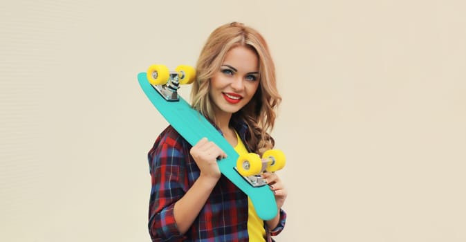 Portrait of stylish young blonde woman posing with skateboard on city street against white wall background