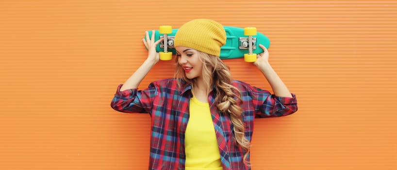 Portrait of stylish happy young blonde woman posing with skateboard in yellow hat, shirt on city street against colorful orange background