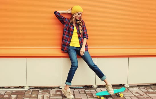 Stylish young blonde woman posing with skateboard in yellow hat, shirt on city street against colorful orange background