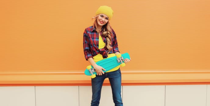 Portrait of stylish happy young blonde woman posing with skateboard in yellow hat, shirt on city street against colorful orange background