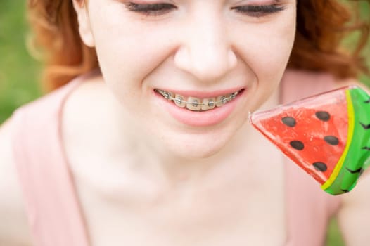 Beautiful young woman with braces on her teeth eats a watermelon-shaped lollipop outdoors