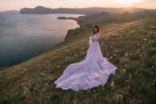 A woman in a long white dress stands on a hill overlooking a body of water. The scene is serene and peaceful, with the woman's dress flowing in the wind