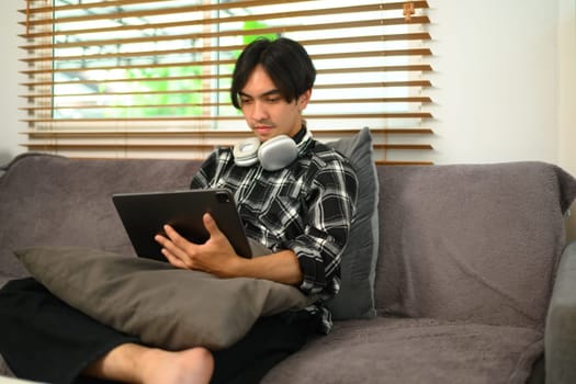 Relaxed young hipster man with headphone using digital tablet on couch at home.