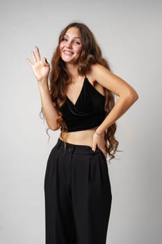 A smiling young woman with long wavy hair raises her index finger as if shes about to express an idea. She wears a black velvet dress and has a playful look on her face. The light grey background provides a neutral backdrop that highlights her figure.