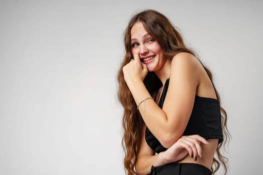 A young woman with long, wavy hair is captured posing expressively with her hand on her cheek and a broad, engaging smile. She is wearing a sleeveless black top which contrasts against a simple, grey backdrop. Her lively expression suggests a moment of genuine happiness or amusement.