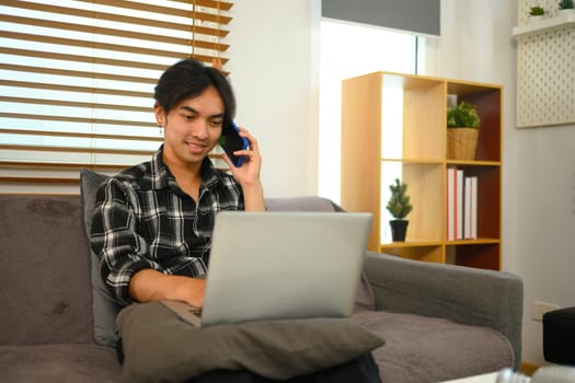 Smiling young man having phone conversation and working with laptop at home.