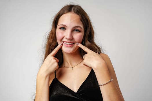 A cheerful young woman with brown hair wearing a black dress is playfully posing against a neutral grey backdrop. She is pointing at her smile, showcasing her happiness, and expressing a positive emotion with sparkling eyes and a beaming expression.