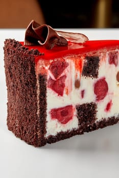 Slice of delicate chocolate biscuit cake filled with whipped sour cream, berry jelly and dessert cherry, topped with vibrant red icing decorated with chocolate petals. Handcrafted sweet treats concept