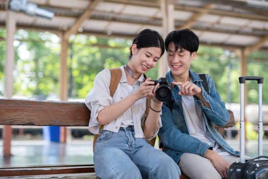 Happy Asian couple holding suitcases and camera preparing to wait for train at train station for vacation trip together..