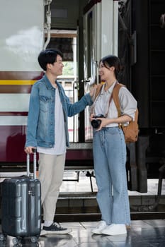 Happy Asian couple holding suitcases and camera preparing to wait for train at train station for vacation trip together..