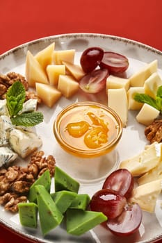 Selection of fine cheeses with crispy walnuts, purple grapes, and glass bowl of clear honey, artistically arranged on plate against red background. Traditional French style appetizer