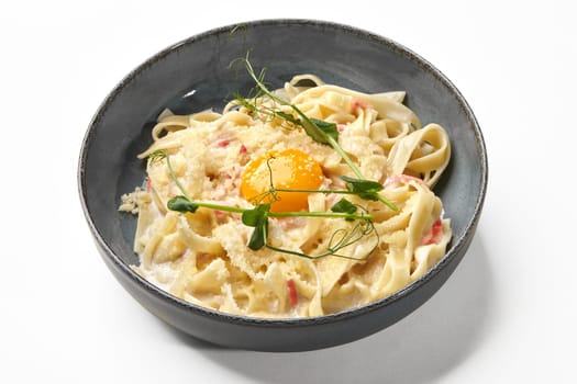 Classic Italian rich creamy fettuccine carbonara with bits of pancetta and egg yolk, garnished with grated parmesan and microgreens, presented in ceramic bowl on white background