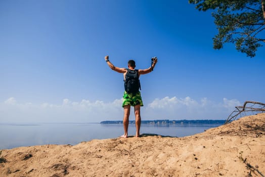 male traveler with their hands up on top with a backpack and a camera over the water