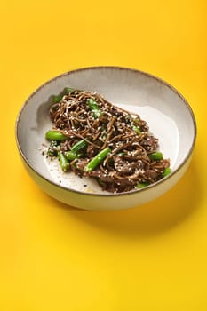 Delicious buckwheat soba noodles with stir-fried beef strips and green beans, sprinkled of black and white sesame seeds served in ceramic bowl against vibrant yellow backdrop. Classic Asian cuisine