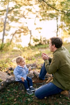 Dad blows on a bouquet of dandelions in his hands on a little girl sitting with her on stumps in the forest. High quality photo