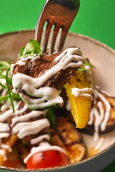 Slices of juicy baked veal and potato drizzled with creamy sauce on fork, poised above plate of warm salad with grilled vegetables and zesty arugula against lively green backdrop