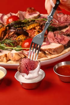 Slice of tender roast beef on fork dipped into creamy sauce, with mixed meat and vegetable grill platter and sauces in background on red surface