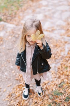 Little girl covers one eye with a yellow leaf while standing in an autumn park. High quality photo