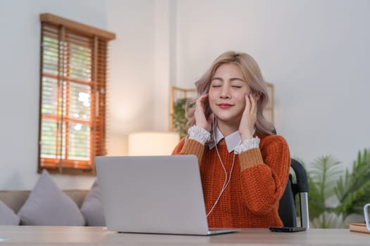 Happy Asian woman listening to music on laptop while sitting on sofa in living room on vacation..