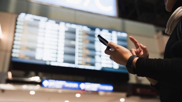 Woman in international airport, using mobile smartphone and checking flight schedule at the flight information board.