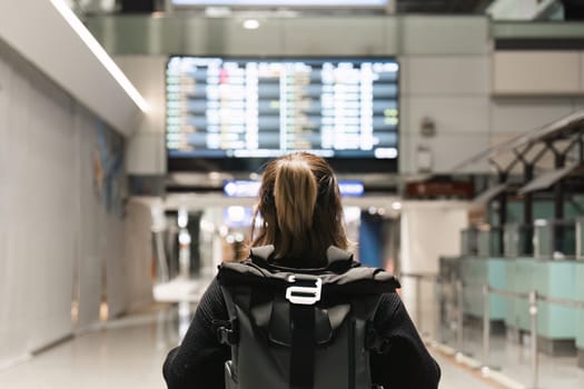 Young woman backpack in international airport terminal looking at the flight schedule information board, checking her flight.