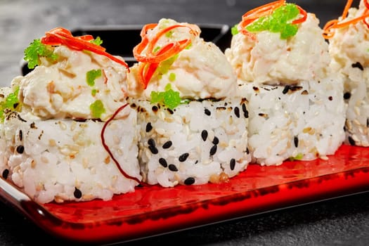Macro shot of elegant sushi roll sprinkled with sesame seeds on ceramic red plate, highlighting creamy toppings with seafood garnished with green tobiko and vegetable shavings. Japanese style delicacy