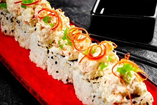 Closeup of sushi rolls topped with cream cheese and shrimp mixture, garnished with spicy chili pepper shavings and green tobiko roe on red plate, on black surface with chopsticks and soy sauce