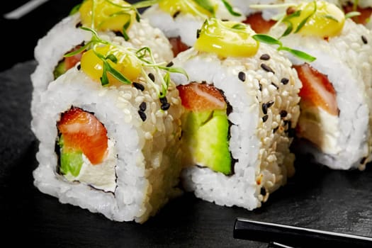 Macro shot showing texture and fresh ingredients of appetizing sushi rolls with salmon, avocado, sesame seeds, drizzled with zesty sauce and sprinkled with greens on dark slate board. Japanese snack