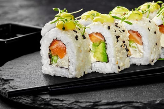 Closeup of appetizing salmon rolls with avocado sprinkled with sesame seeds topped with drops of savory sauce and fresh microgreens, presented on black slate serving board. Japanese snack concept