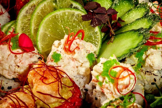 Macro shot of colorful sushi rolls platter garnished with green tobiko, togarashi threads and pepper shavings, accompanied with sliced cucumber and lime. Japanese fusion cuisine