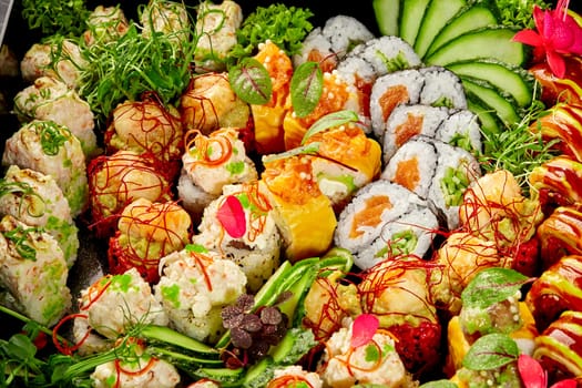 Colorful assorted sushi platter with traditional Japanese rolls with seafood and vegetables garnished with slices of fresh cucumber, lime, microgreens and flowers, presented on dark background