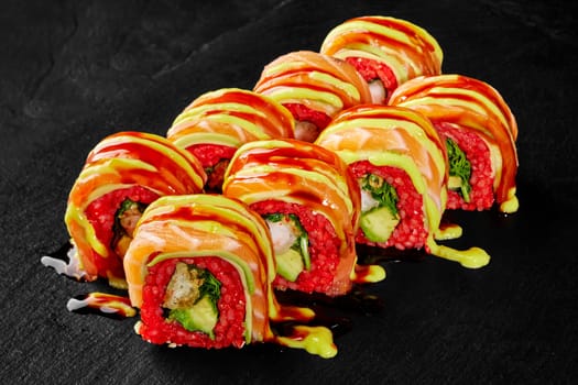 Spicy colorful red rice uramaki rolls filled with shrimp tempura, avocado and lettuce, topped with fresh salmon, drizzled with unagi sauce and mayo, served on black slate board. Japanese cuisine