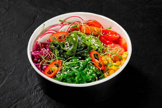 Colorful vegan poke bowl with sliced avocado, radishes, tomatoes, hiyashi wakame, corn, greens and hot chili peppers sprinkled with sesame seeds, presented in cardboard cup on dark textured surface