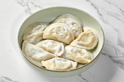 Delicate sweet Polish pierogi filled with fresh juicy berries or fruits served in rustic bowl, generously sprinkled with powdered sugar, on clean marble background